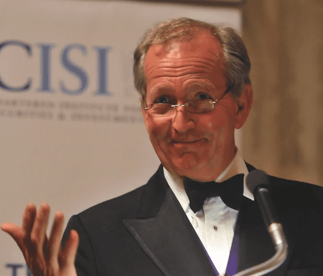 A man in a tuxedo speaks at a microphone with a backdrop.